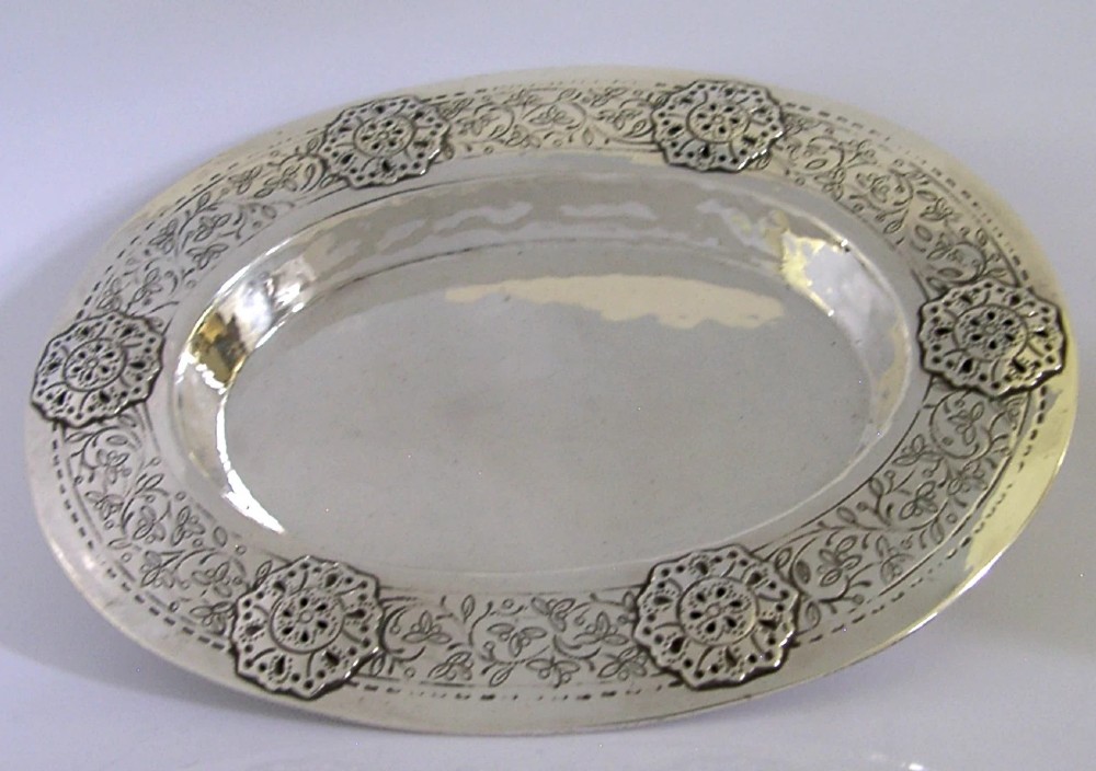 An oval antique sterling silver dish featuring a planished centre and a wide rim decorated with chased floral rosettes and leaf motifs. View VI