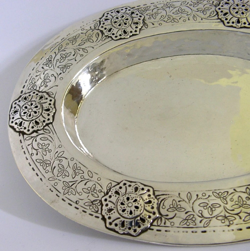 An oval antique sterling silver dish featuring a planished centre and a wide rim decorated with chased floral rosettes and leaf motifs. View VII