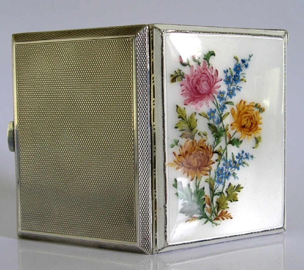 A square sterling silver hinged cigarette case showing an engine-turned textured exterior on one side and a white enamelled panel with hand-painted yellow and pink chrysanthemums on the other.