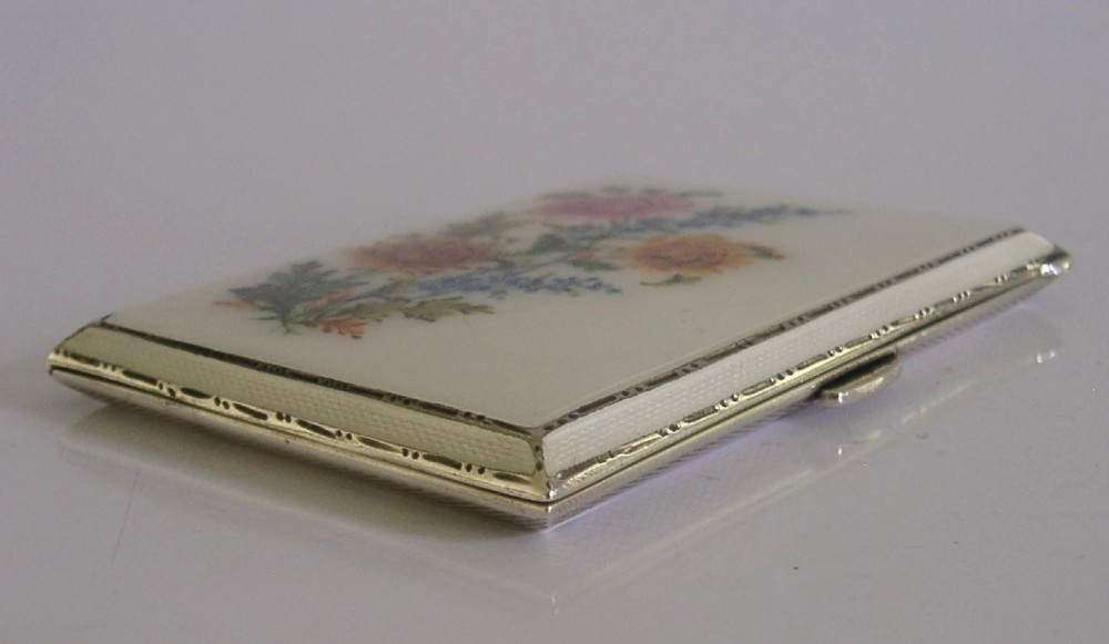 A square sterling silver hinged cigarette case showing an engine-turned textured exterior on one side and a white enamelled panel with hand-painted yellow and pink chrysanthemums on the other. View IX