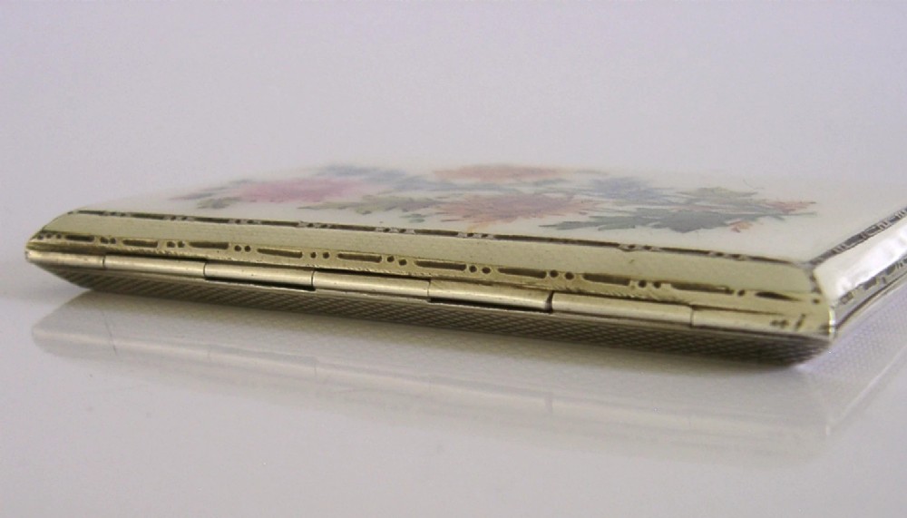 A square sterling silver hinged cigarette case showing an engine-turned textured exterior on one side and a white enamelled panel with hand-painted yellow and pink chrysanthemums on the other. View X
