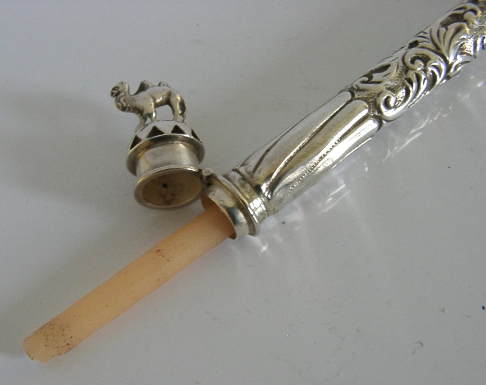 An elongated cylindrical silver Victorian wax holder featuring scrollwork engraving and a camel figure at the terminal. View IV