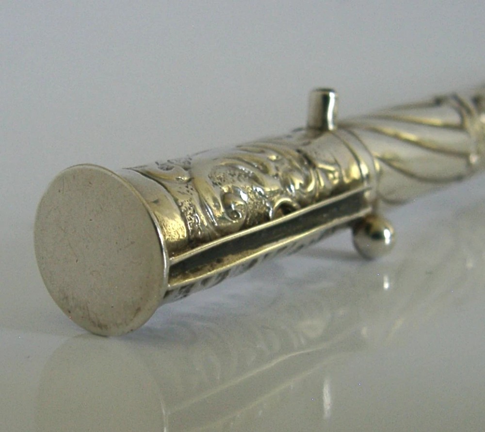 An elongated cylindrical silver Victorian wax holder featuring scrollwork engraving and a camel figure at the terminal. View VI