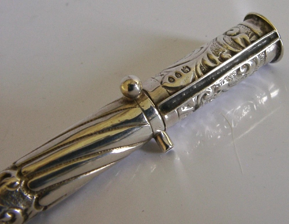 An elongated cylindrical silver Victorian wax holder featuring scrollwork engraving and a camel figure at the terminal. View IX