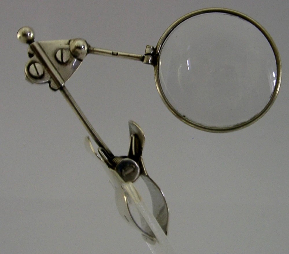 A sterling silver thumb magnifier glass with an adjustable arm clip rested inside a black velvet and silk lined presentation box. View V