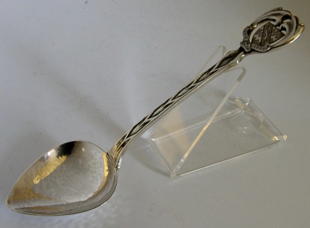 An Art Deco sterling silver spoon featuring an openwork coat of arms handle terminal, a geometric stem, and a polished teardrop bowl. View VI