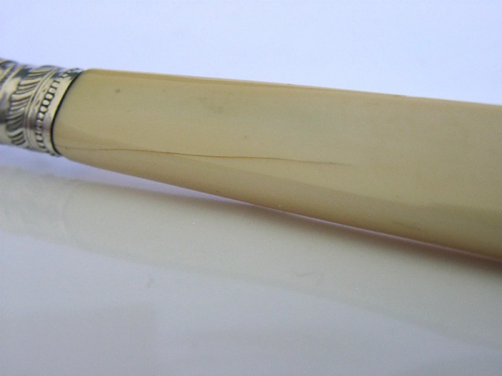 An Edwardian sterling silver cheese knife featuring a curved blade with two terminal prongs, a decorative chased collar, and a smooth cream handle. View VI