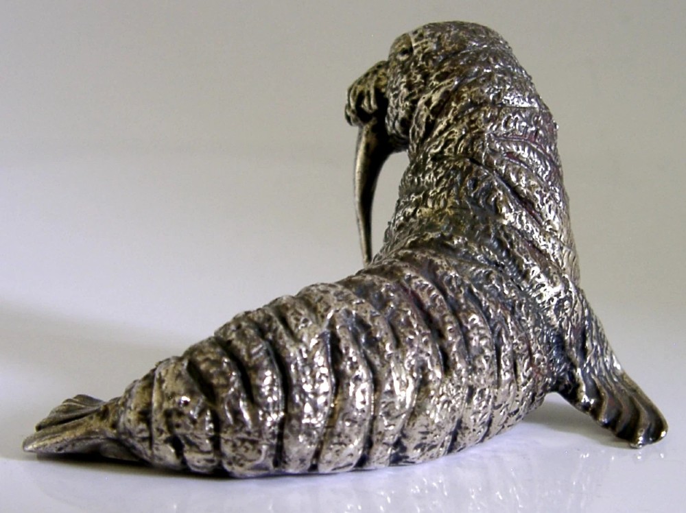 A detailed cast silver walrus figurine shown in profile with long downward tusks and textured skin. View VII