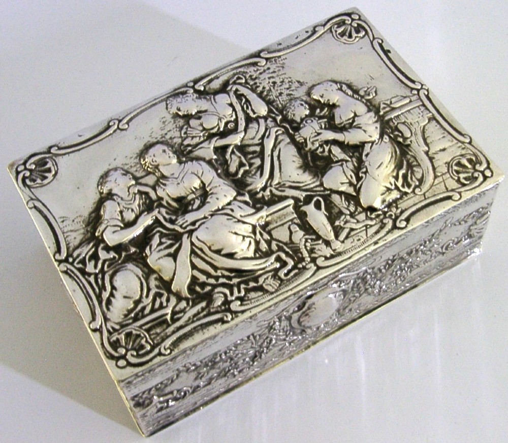 A rectangular silver box featuring embossed classical figures on the hinged lid and foliate scrollwork along the sides.
