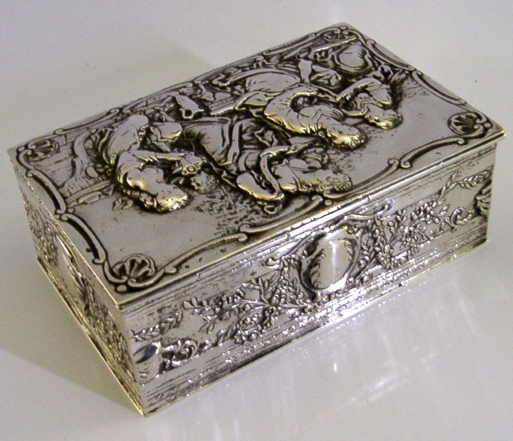 A rectangular silver box featuring embossed classical figures on the hinged lid and foliate scrollwork along the sides. View V