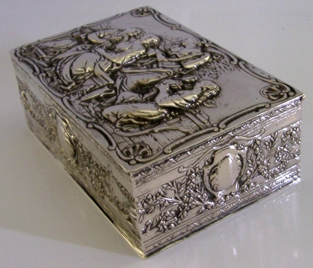 A rectangular silver box featuring embossed classical figures on the hinged lid and foliate scrollwork along the sides. View VII