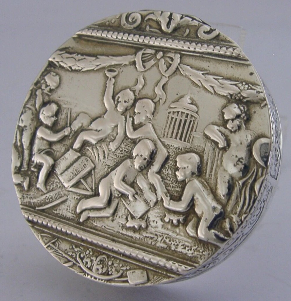 A circular antique silver trinket box featuring embossed relief depicting putti playing beneath floral garlands.