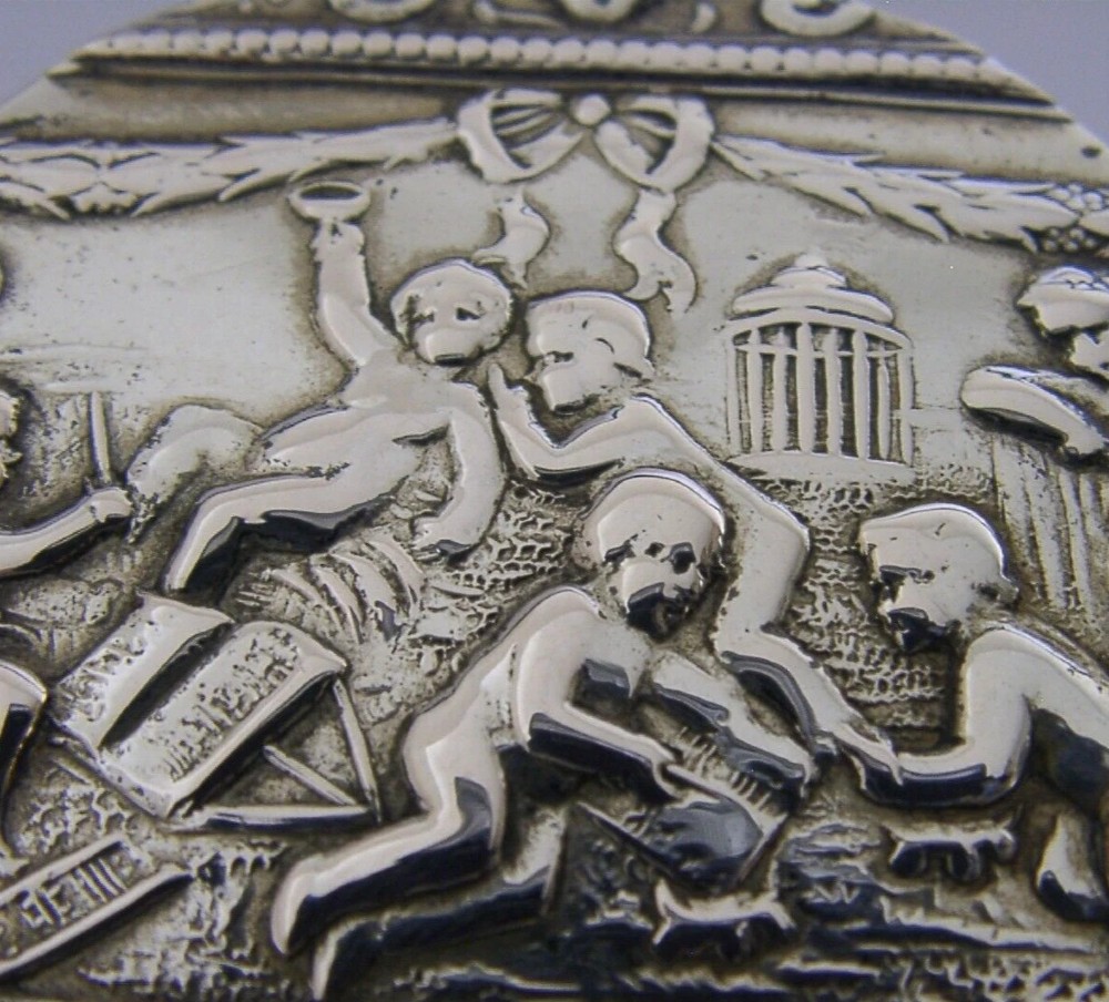 A circular antique silver trinket box featuring embossed relief depicting putti playing beneath floral garlands. View II