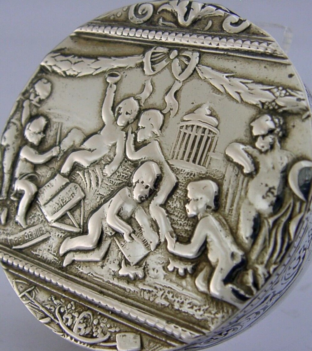 A circular antique silver trinket box featuring embossed relief depicting putti playing beneath floral garlands. View III