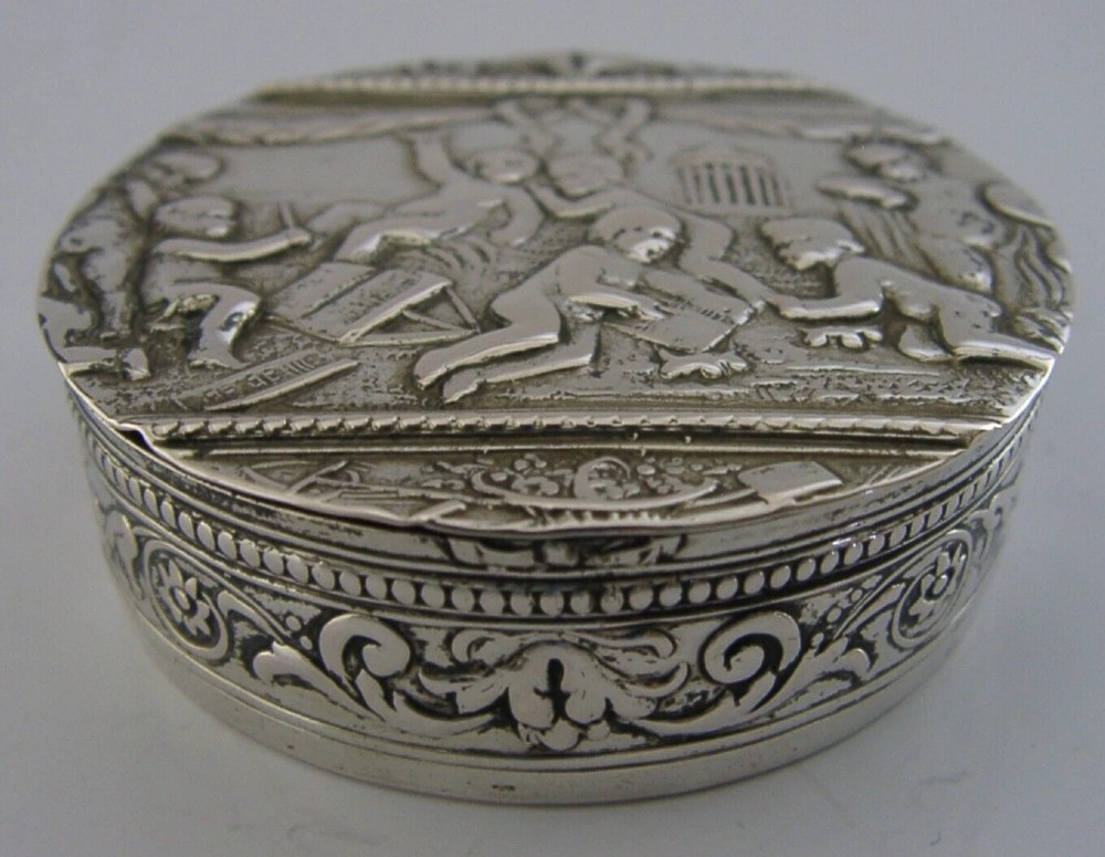 A circular antique silver trinket box featuring embossed relief depicting putti playing beneath floral garlands. View VI