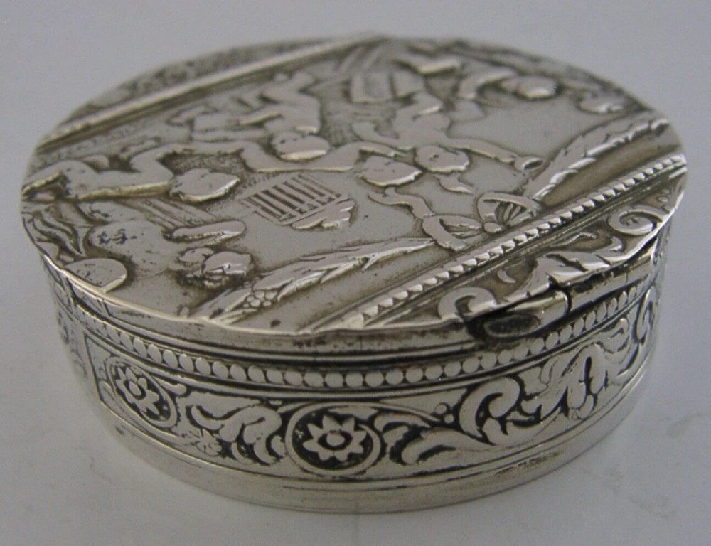 A circular antique silver trinket box featuring embossed relief depicting putti playing beneath floral garlands. View VII