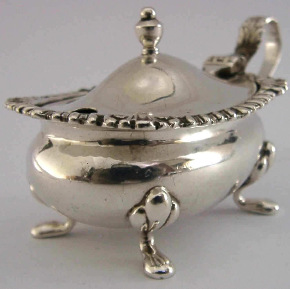 A solid sterling silver mustard pot featuring a domed hinged lid with a finial, decorative scalloped rim, scroll handle, and four pad feet.