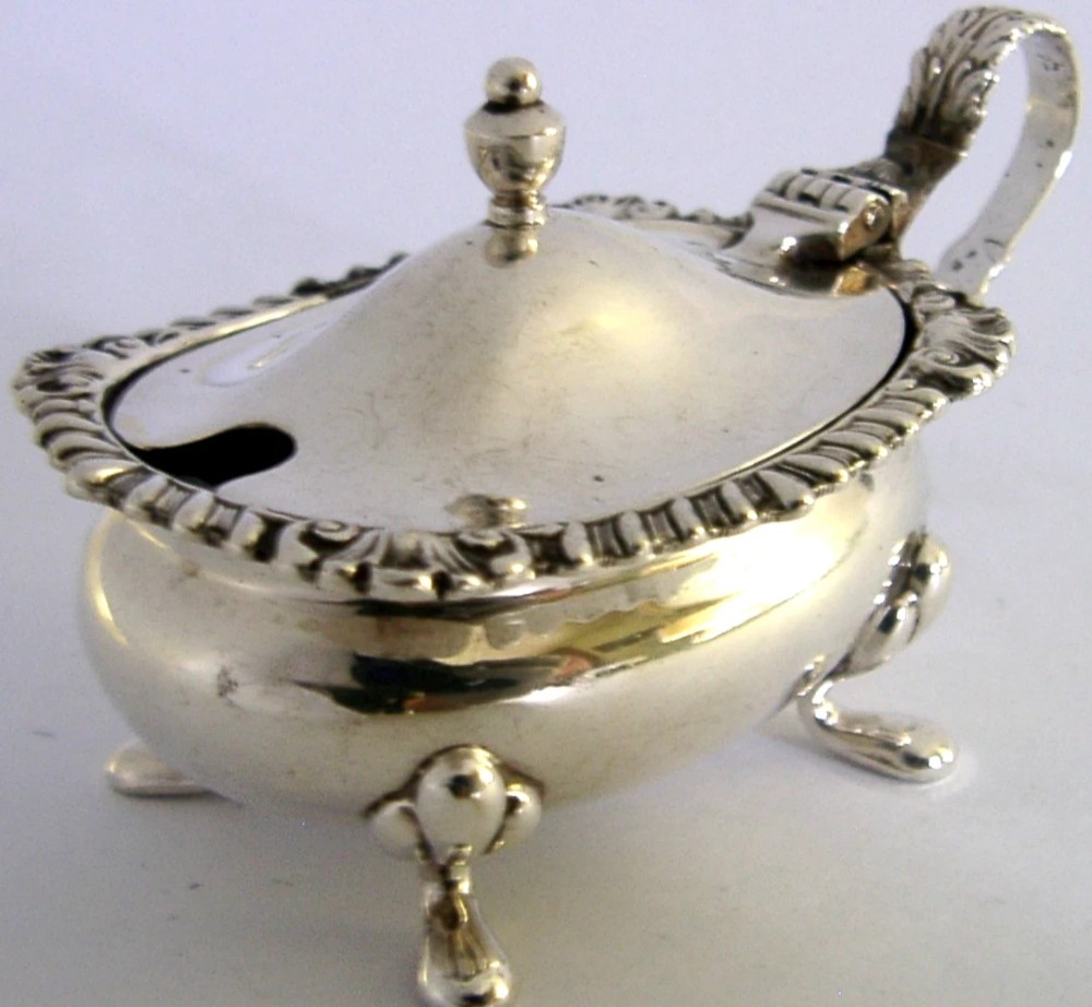 A solid sterling silver mustard pot featuring a domed hinged lid with a finial, decorative scalloped rim, scroll handle, and four pad feet. View II