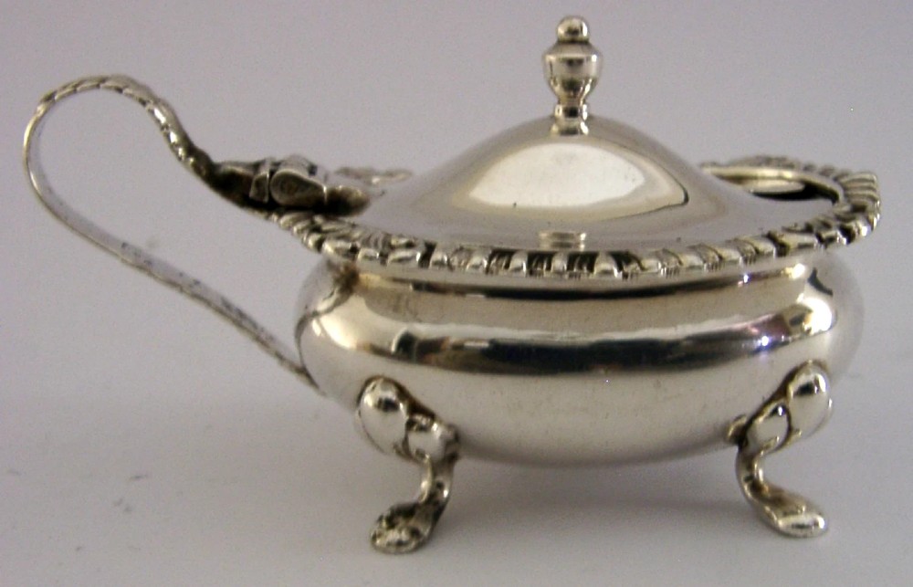 A solid sterling silver mustard pot featuring a domed hinged lid with a finial, decorative scalloped rim, scroll handle, and four pad feet. View III