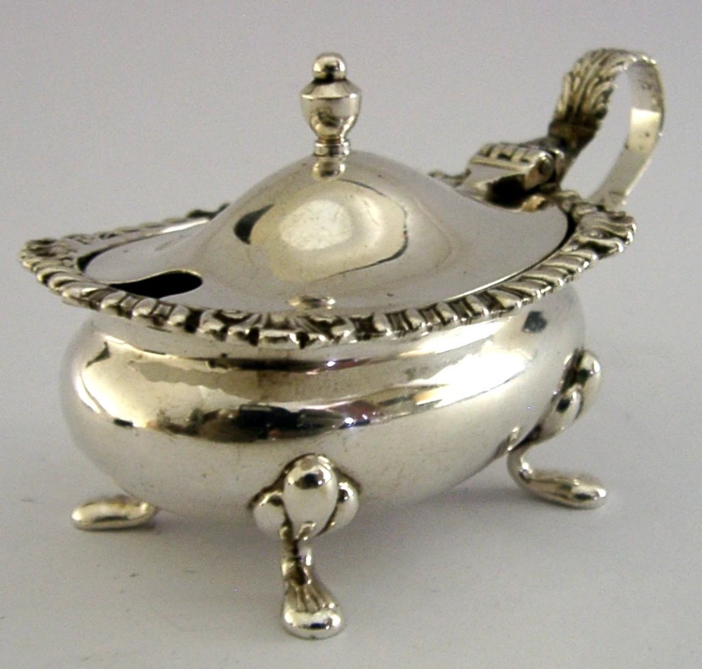 A solid sterling silver mustard pot featuring a domed hinged lid with a finial, decorative scalloped rim, scroll handle, and four pad feet. View IV