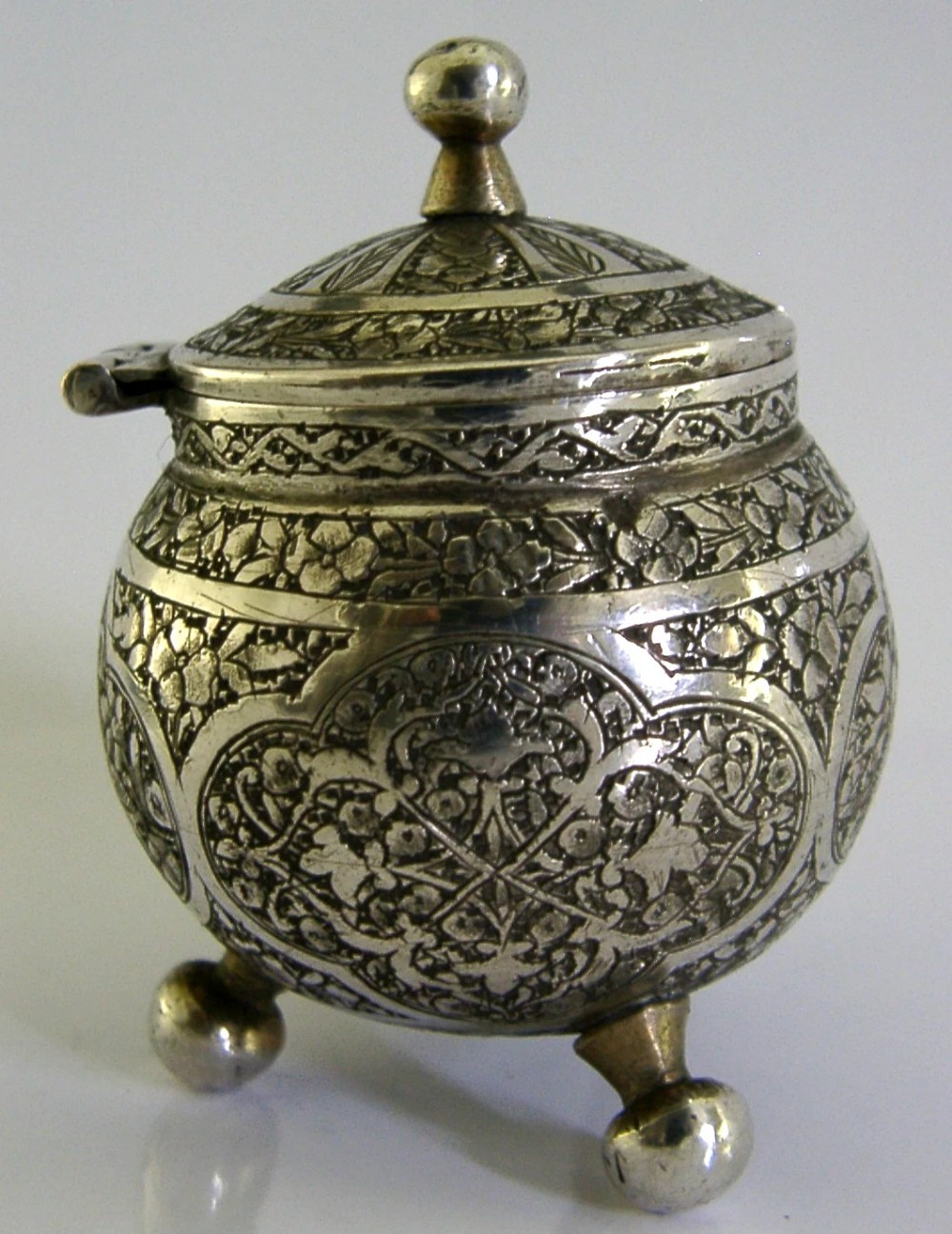 An antique hand-engraved solid silver mustard pot featuring arabesque foliage patterns, a hinged lid with finial, a spoon aperture, and three ball feet. View V
