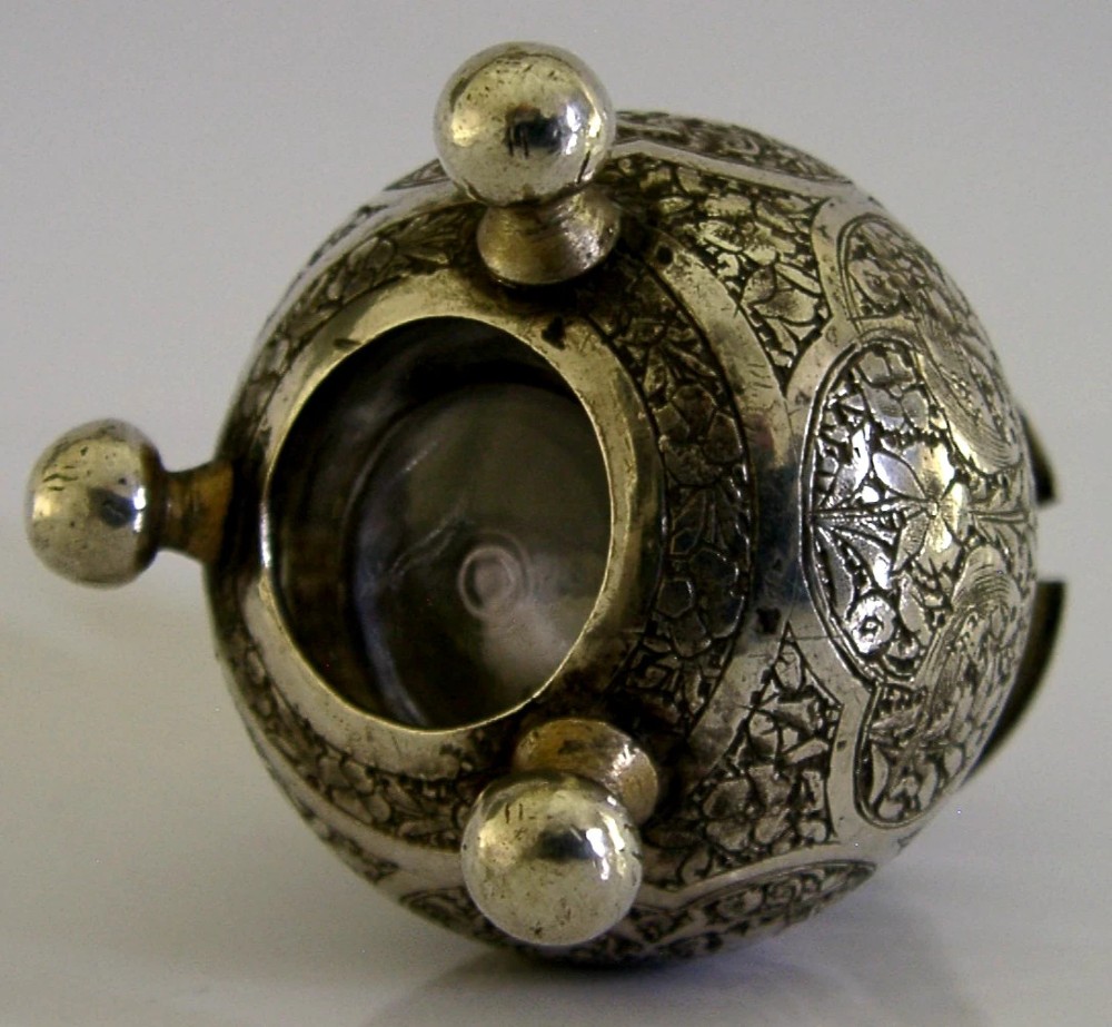 An antique hand-engraved solid silver mustard pot featuring arabesque foliage patterns, a hinged lid with finial, a spoon aperture, and three ball feet. View IX