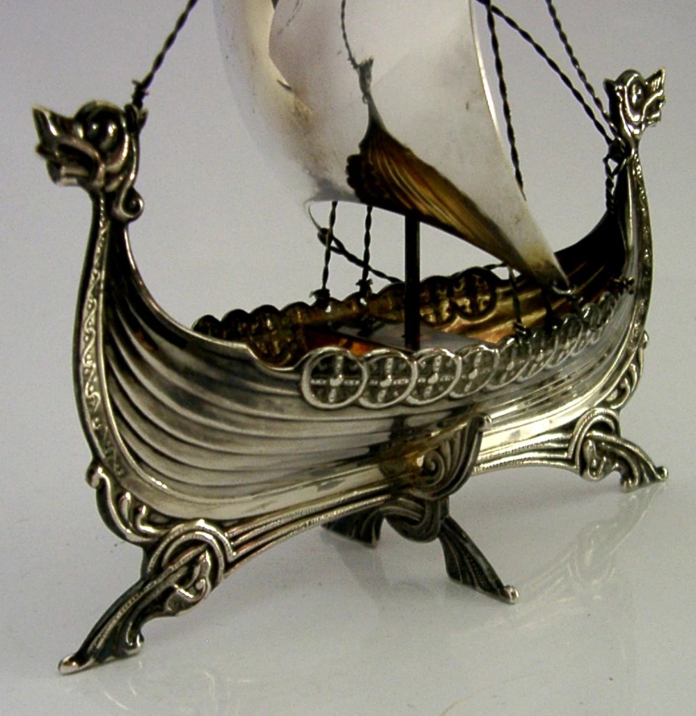 A detailed solid silver model of a Viking longship featuring a dragon head prow, raised sail, pennant flag, and miniature shields along the gunwale. View III