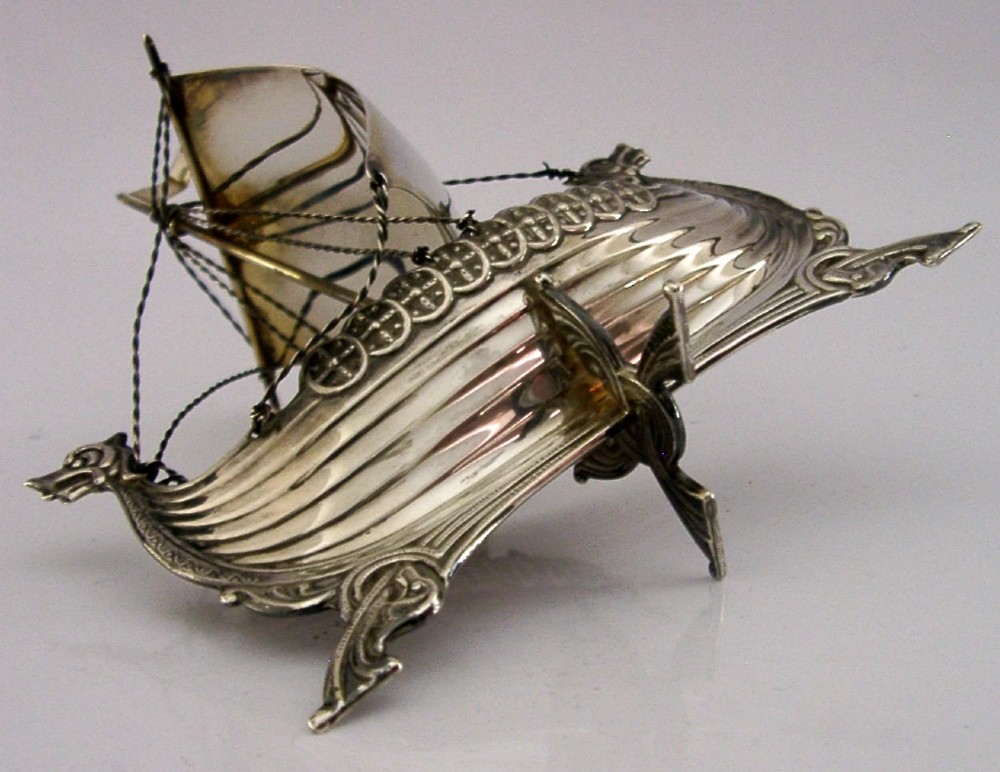 A detailed solid silver model of a Viking longship featuring a dragon head prow, raised sail, pennant flag, and miniature shields along the gunwale. View IV