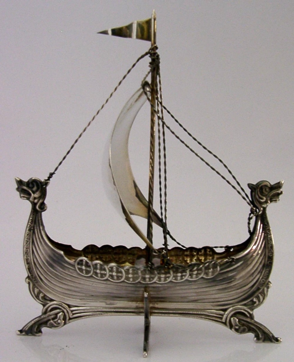 A detailed solid silver model of a Viking longship featuring a dragon head prow, raised sail, pennant flag, and miniature shields along the gunwale. View VIII