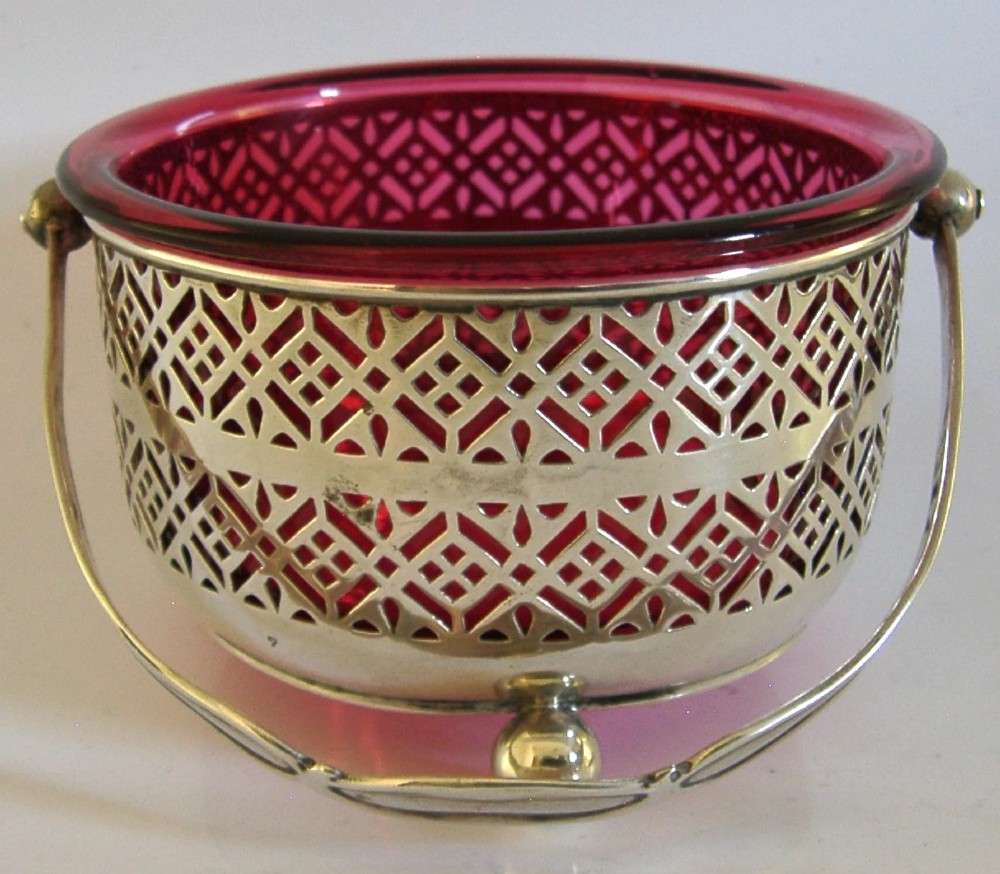 An Edwardian sterling silver pierced basket featuring a lattice geometric pattern, a swing handle, and a fitted cranberry red glass liner.
