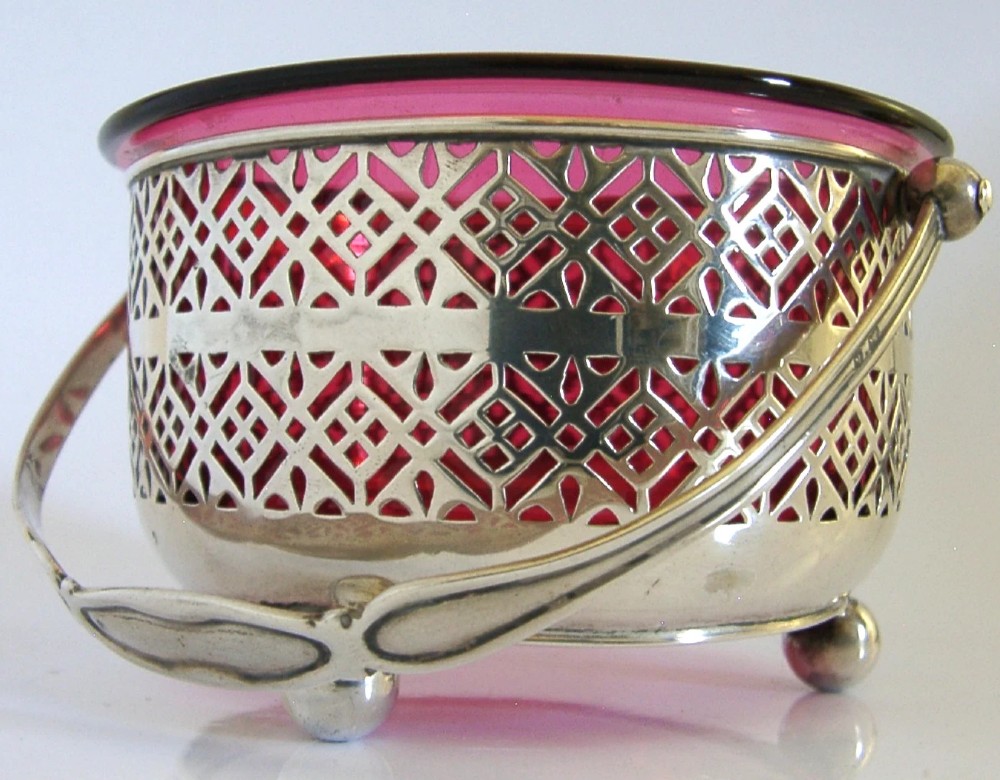 An Edwardian sterling silver pierced basket featuring a lattice geometric pattern, a swing handle, and a fitted cranberry red glass liner. View V