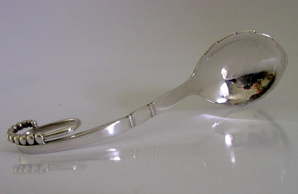 A sterling silver Georg Jensen spoon featuring a curved loop handle detailed with beaded ornamentation and a shallow oval bowl.