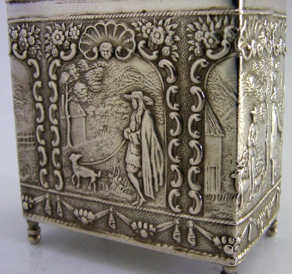 A rectangular solid silver tea caddy featuring relief-embossed pastoral scenes, turned feet, and a finial lid. View II