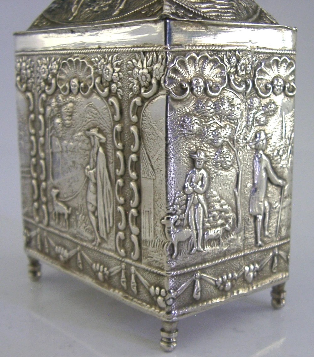 A rectangular solid silver tea caddy featuring relief-embossed pastoral scenes, turned feet, and a finial lid. View IV