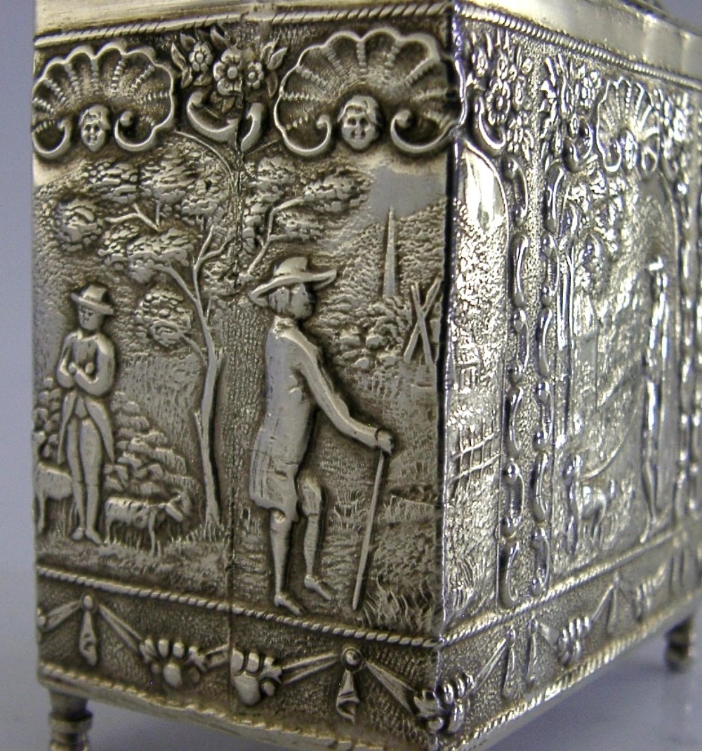A rectangular solid silver tea caddy featuring relief-embossed pastoral scenes, turned feet, and a finial lid. View V