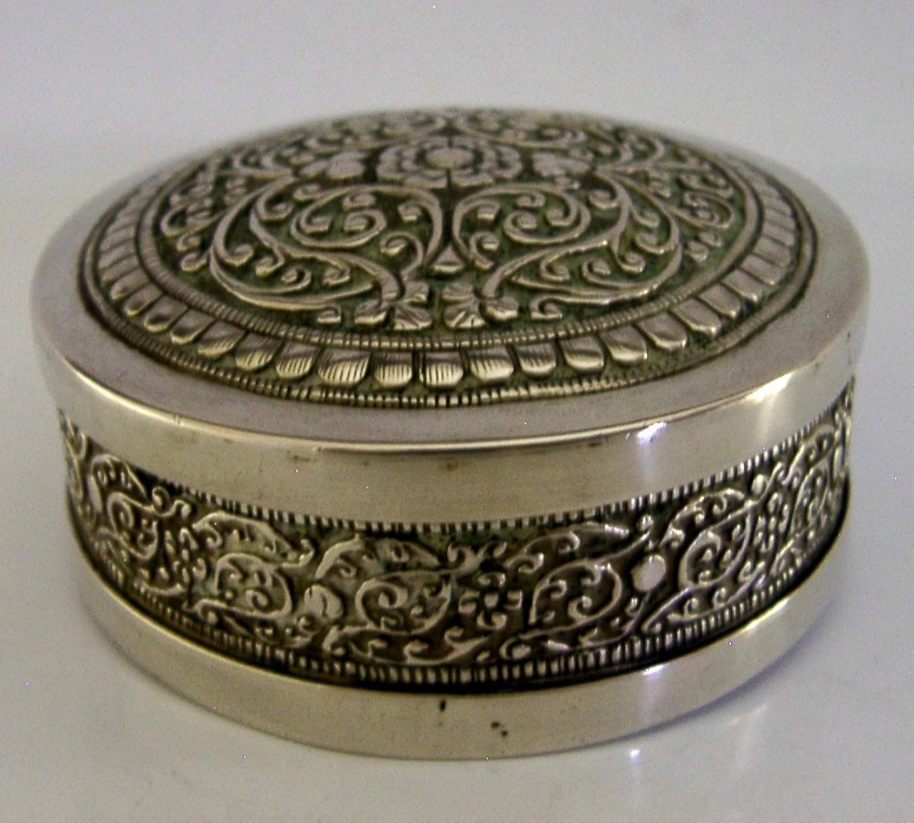 A circular antique silver trinket box with a lidded top featuring dense relief floral scrollwork and an oxidised background. View III