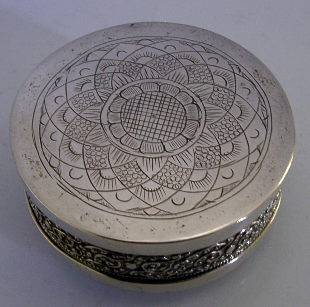 A circular antique silver trinket box with a lidded top featuring dense relief floral scrollwork and an oxidised background. View V
