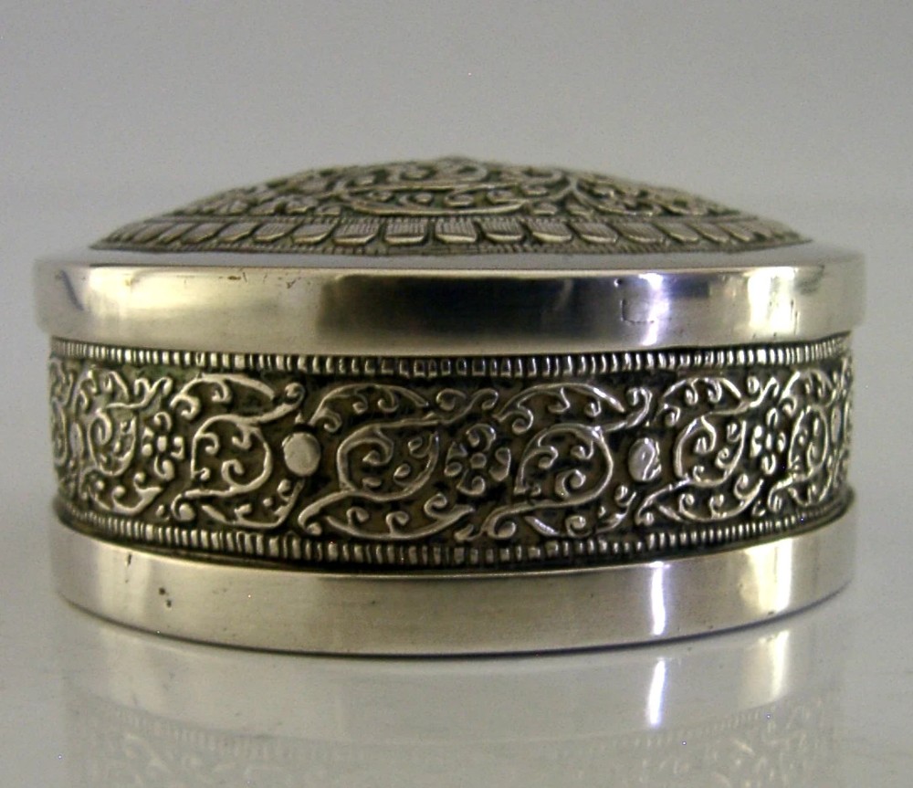 A circular antique solid silver box with lid, decorated with dense foliate repousse motifs and floral chasing. View II