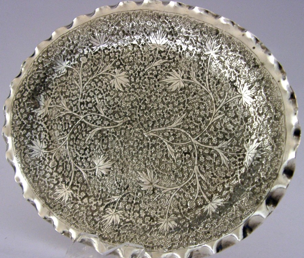 An oval solid silver dish featuring dense chased leaf patterns and a scalloped fluted border.