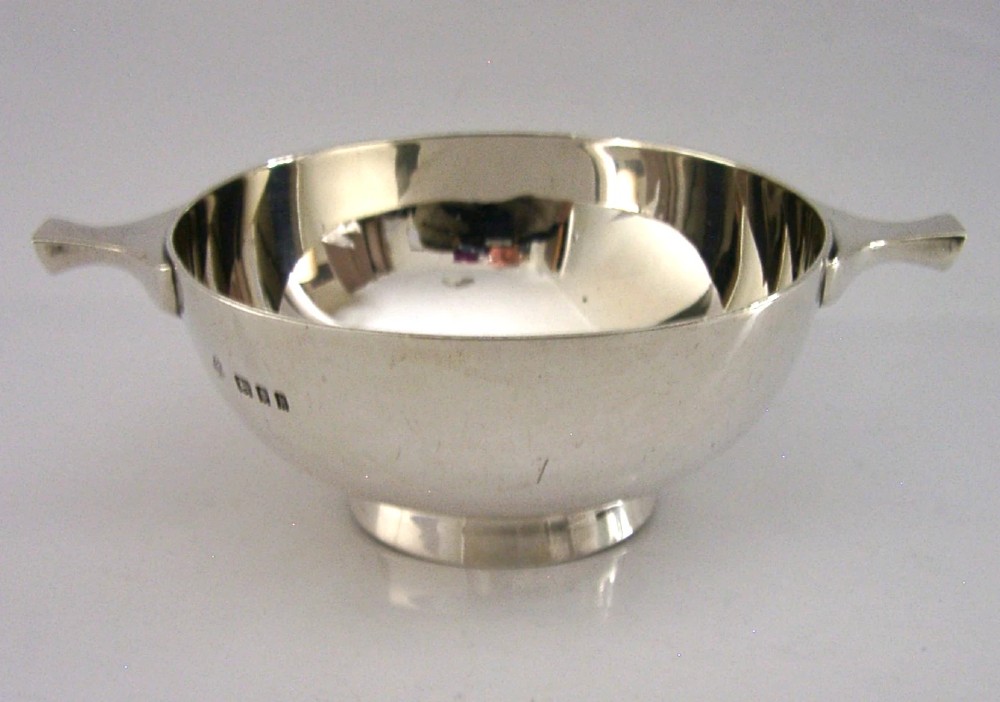 A polished sterling silver shallow drinking cup featuring two flat side handles and British hallmarks stamped on the exterior wall.