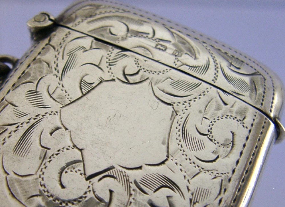 An antique sterling silver vesta case featuring hand-engraved foliate scrollwork, a central shield cartouche, a hinged lid, and a side suspension ring. View IV