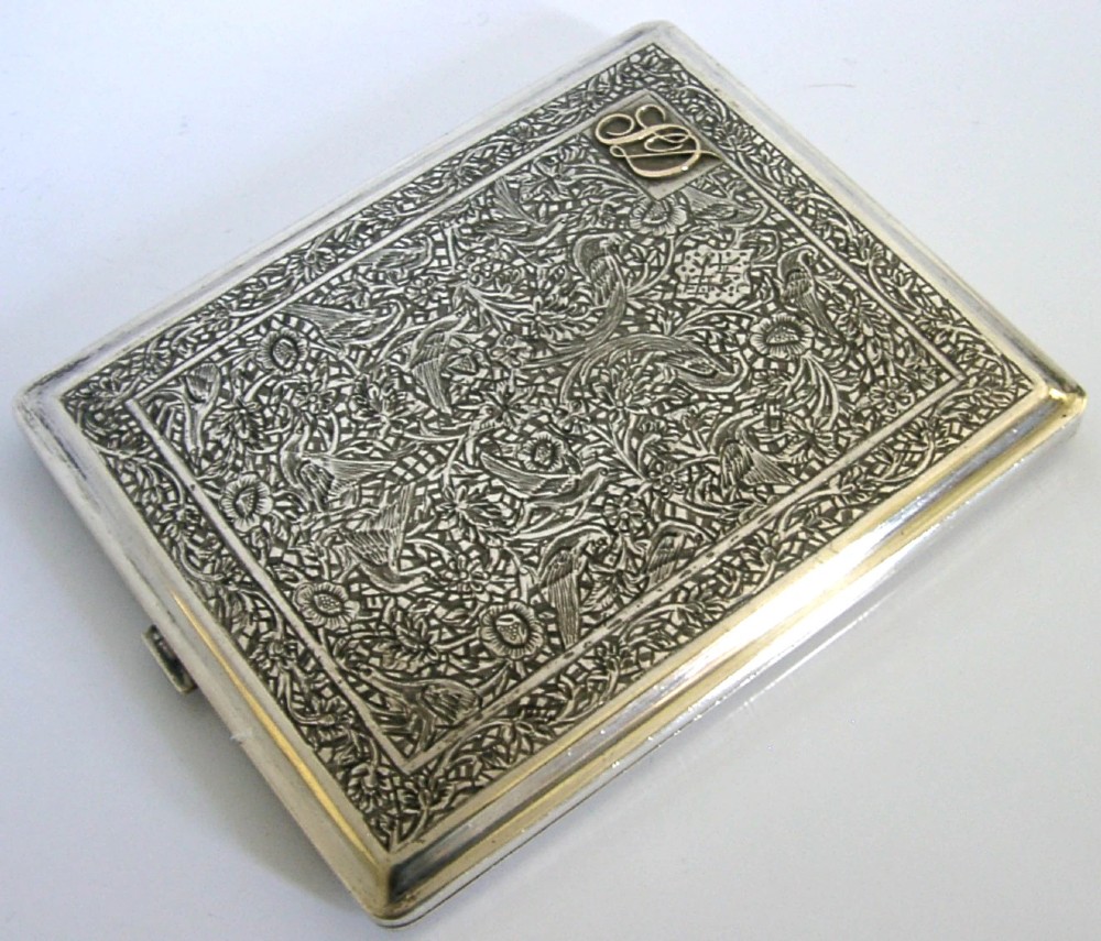 A rectangular solid silver cigarette case featuring dense hand-engraved bird and foliate patterns with a gold monogram in the upper corner. View IV