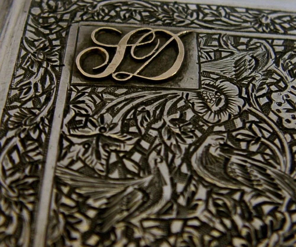 A rectangular solid silver cigarette case featuring dense hand-engraved bird and foliate patterns with a gold monogram in the upper corner. View V