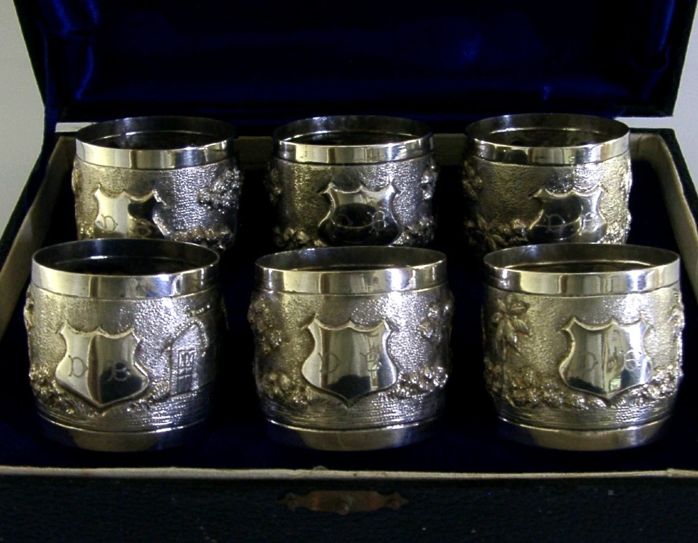 A set of six silver napkin rings with repousse landscape relief patterns and shield cartouches, arranged inside a blue velvet-lined fitted box.