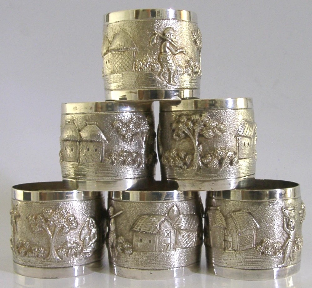 A set of six silver napkin rings with repousse landscape relief patterns and shield cartouches, arranged inside a blue velvet-lined fitted box. View II