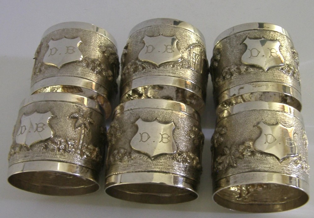 A set of six silver napkin rings with repousse landscape relief patterns and shield cartouches, arranged inside a blue velvet-lined fitted box. View IV