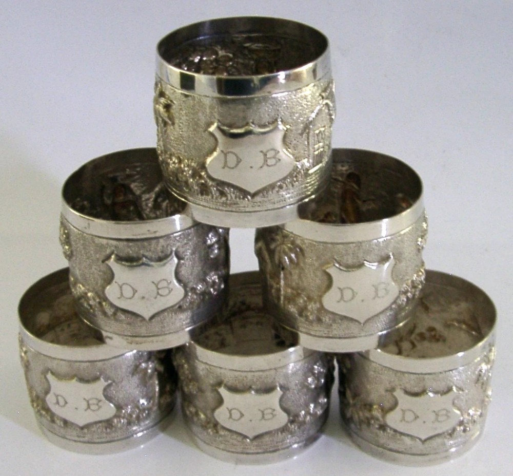 A set of six silver napkin rings with repousse landscape relief patterns and shield cartouches, arranged inside a blue velvet-lined fitted box. View V