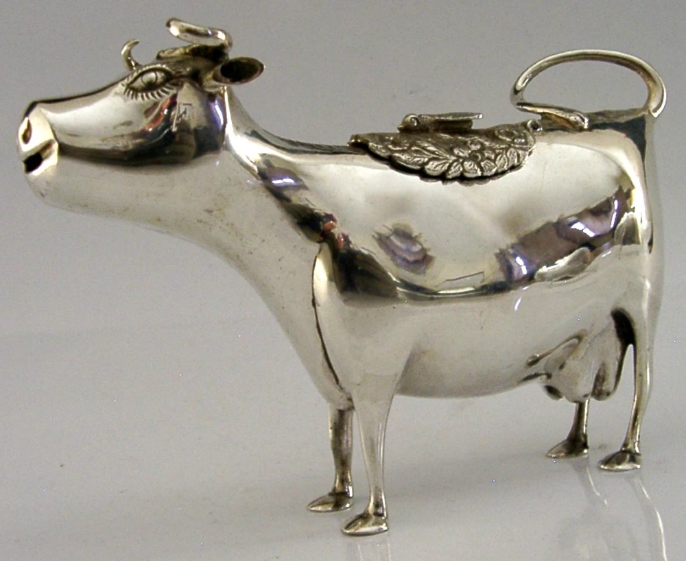 A sterling silver novelty cow creamer featuring a hollow cast body, a hinged lid decorated with floral motifs on the back, and a looped tail handle.