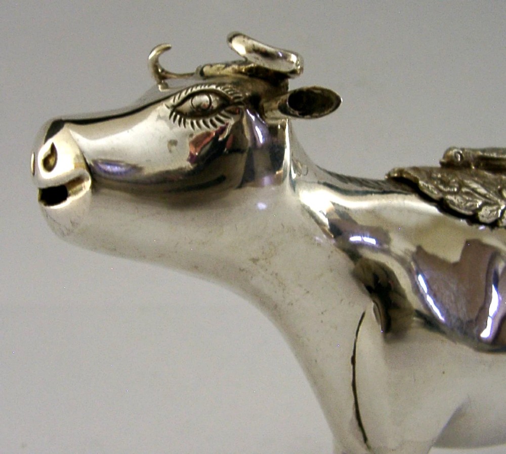 A sterling silver novelty cow creamer featuring a hollow cast body, a hinged lid decorated with floral motifs on the back, and a looped tail handle. View II