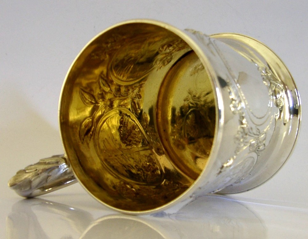 A Victorian sterling silver tankard mug featuring detailed relief floral decoration, a landscape roundel, a scroll handle, and a gilt interior. View IV